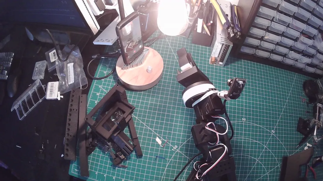 Bench camera still: an SO-101 robot arm on a cutting mat with a black tool locked on its wrist and a rubber-banded wrist camera, with loose parts on the mat