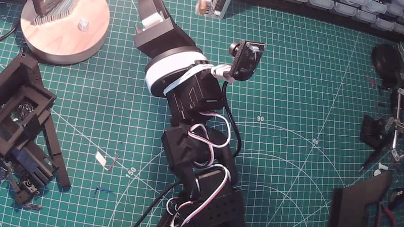Bench camera still: an SO-101 robot arm on a cutting mat with a black tool locked on its wrist and a rubber-banded wrist camera, with loose parts on the mat
